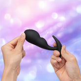 Two hands hold and stretch a black B-Vibe Vibrating P-Spot Snug Plug RC Large by B-Vibe against a blurred, pastel purple and blue background with bokeh lighting effects.